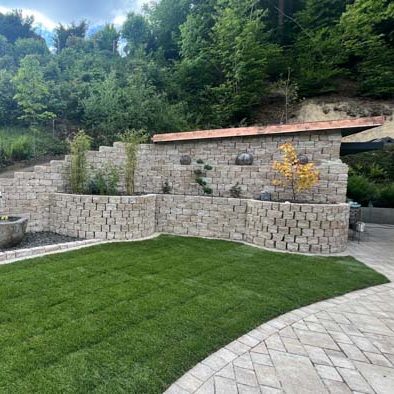 Mauer aus hellen Steinen mit Pflanzen, umgeben von einem gepflasterten Bereich.