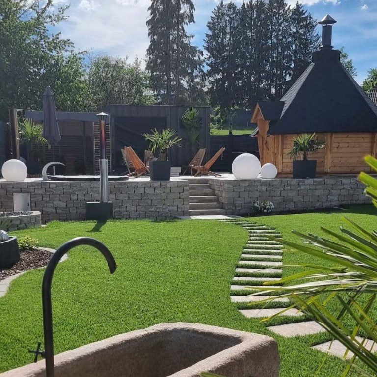 Garten mit Rasen, Steinstufen und Holzhütte hinter schattenspendenden Bäumen.